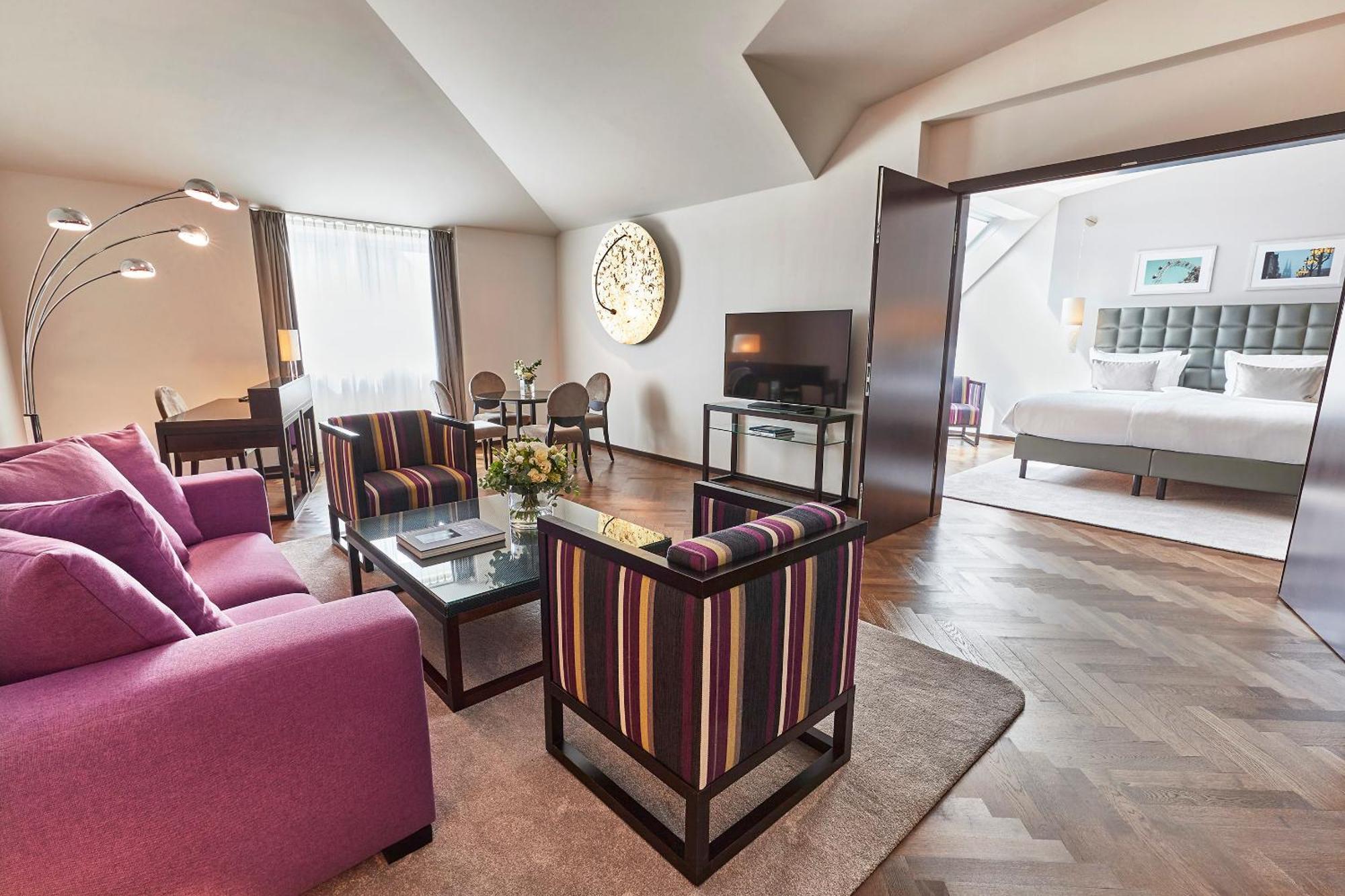 Steigenberger Hotel Herrenhof Wina Bagian luar foto The photo shows a modern, stylish living space that appears to be part of a hotel suite. The room features a cozy seating area with a purple sofa and two striped chairs around a glass coffee table. A flat-screen TV is mounted on the wall. Large windo